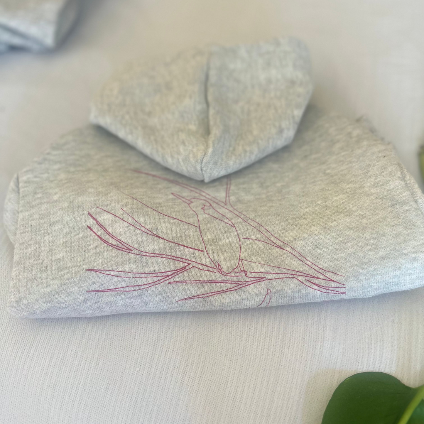 A neatly folded flat lay of the rose-printed ‘Charlie and the Bird’ hoodie in size small, showcasing the intricate bird design prominently. The arms are tucked in, and the hoodie lies flat on a white background with a subtle pinstripe texture, adding a touch of elegance. The corner of another hoodie is partially visible in the left, while a leaf is subtly cropped in the right corner, enhancing the natural feel of the image