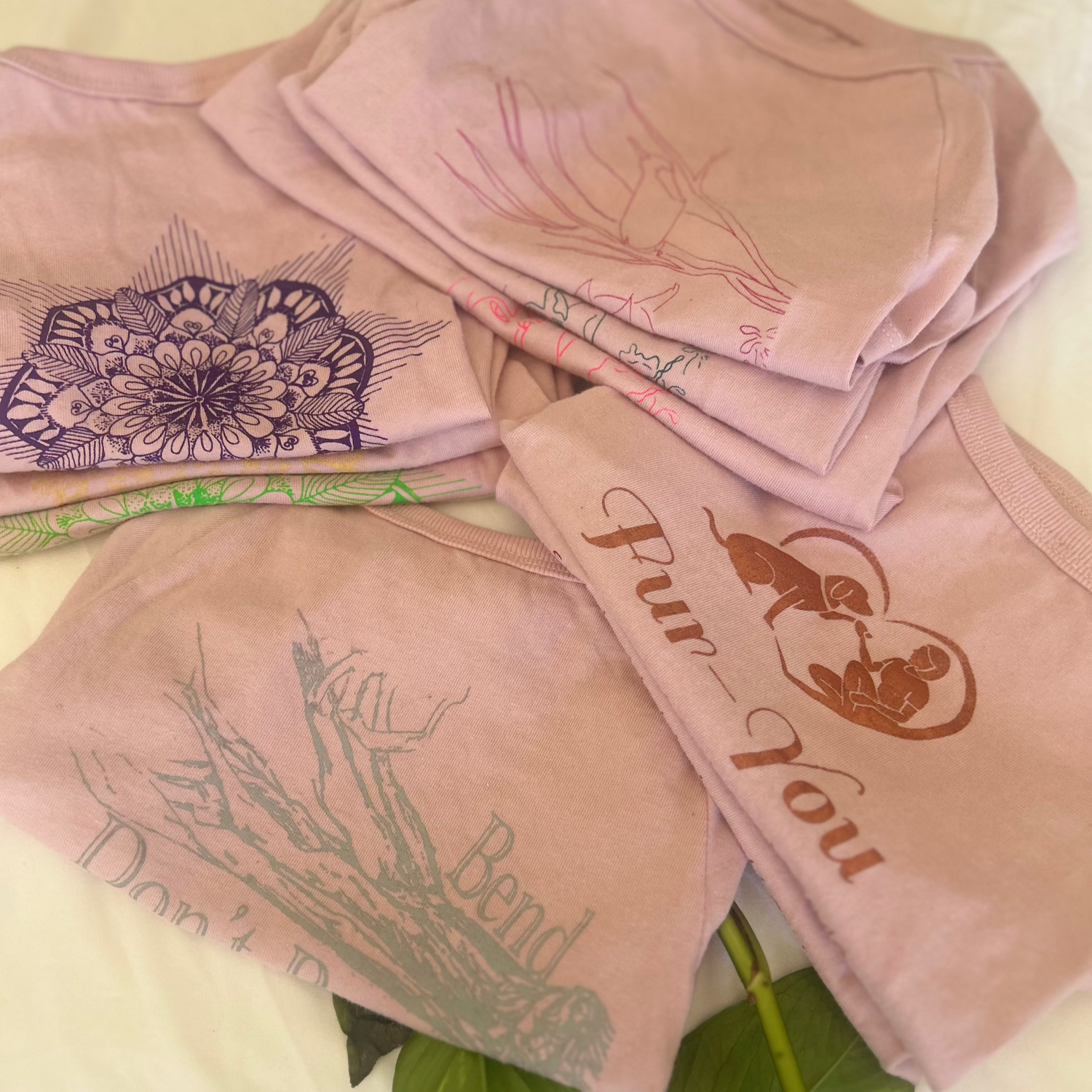 A neatly folded collection of Fur-You’s sustainable dog tees in dusty pink, showcasing intricate hand-screen-printed designs. The top left stack features the Mandala print in glow green, gold, and purple, while the bottom left displays the ‘Bend Don’t Break’ print in grey, accented by natural vine leaves. A Fur-You logo tee stack sits on the right, topped with the Copper print. At the centre, a stack of ‘Charlie and the Bird’ tees in glow pink, green, and rose adds an artistic touch. Ethically made from pre