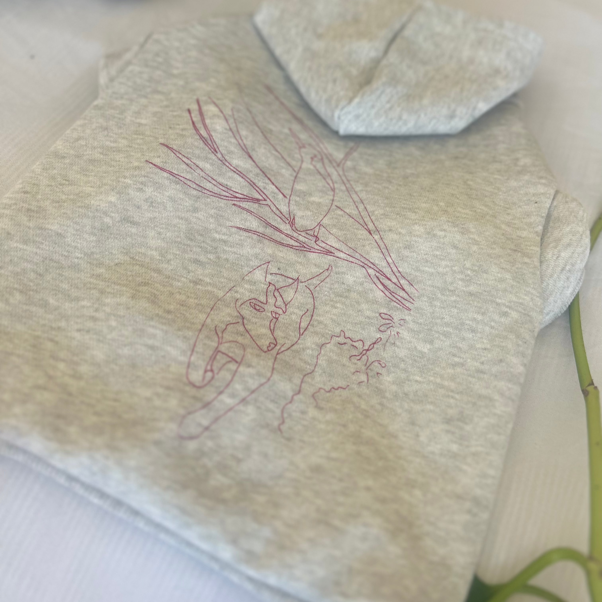 A flat lay of the grey marle ‘Charlie and the Bird’ hoodie, showcasing the full back print in elegant rose ink. The hoodie is neatly folded, with arms tucked underneath, resting on a white surface featuring a subtle pinstripe pattern. A vine and leaf partially frame the lower left corner, enhancing the serene and nature-inspired feel. This image highlights the sustainable craftsmanship and artistic design that defines Fur-You’s petwear collection