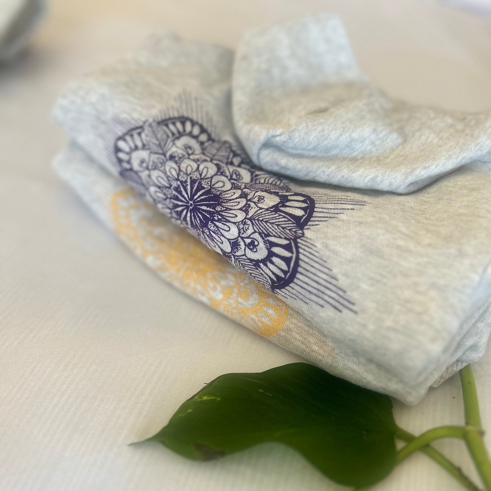 The purple hoodie and gold lustre is beautifully folded on a white background, accompanied by vine leaves beneath. The subtle placement of another hoodie in the left corner adds a layered, dynamic feel. This visual showcases the hoodie’s luxurious feel while reinforcing the brand’s commitment to using sustainable materials and eco-friendly practices.