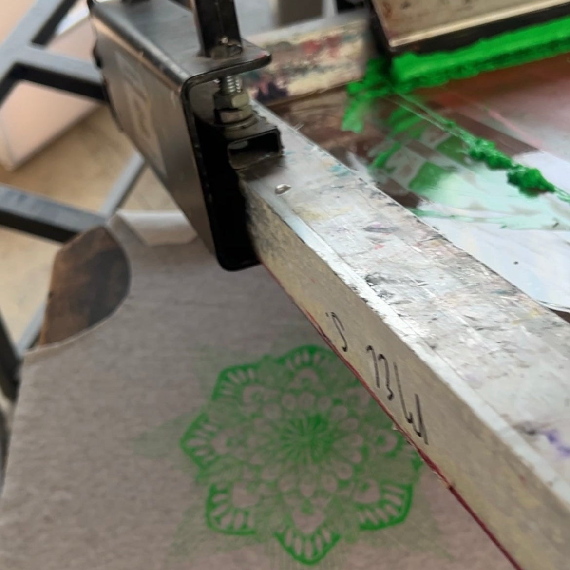 Screen slightly off to the right with squeegee and glow green ink just printed. Below, a hoodie fabric panel with the glow green mandala print. Captures the hands-on, artisanal process