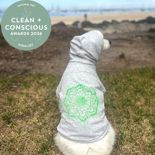 Nominated finalist for natural pet accessory in clean + concious awards 2026 rosette in top left corner of image for a dog sits on the grass leading to the sand and bayside of Melbourne in the background. Full back view of the hoodie with the hood up shows the relaxed, comfortable fit that’s perfect for outdoor adventures. The eco-friendly glow green screen print of the Mandala subtly adds to the overall look, enhancing the hoodie’s sustainable yet stylish appeal.