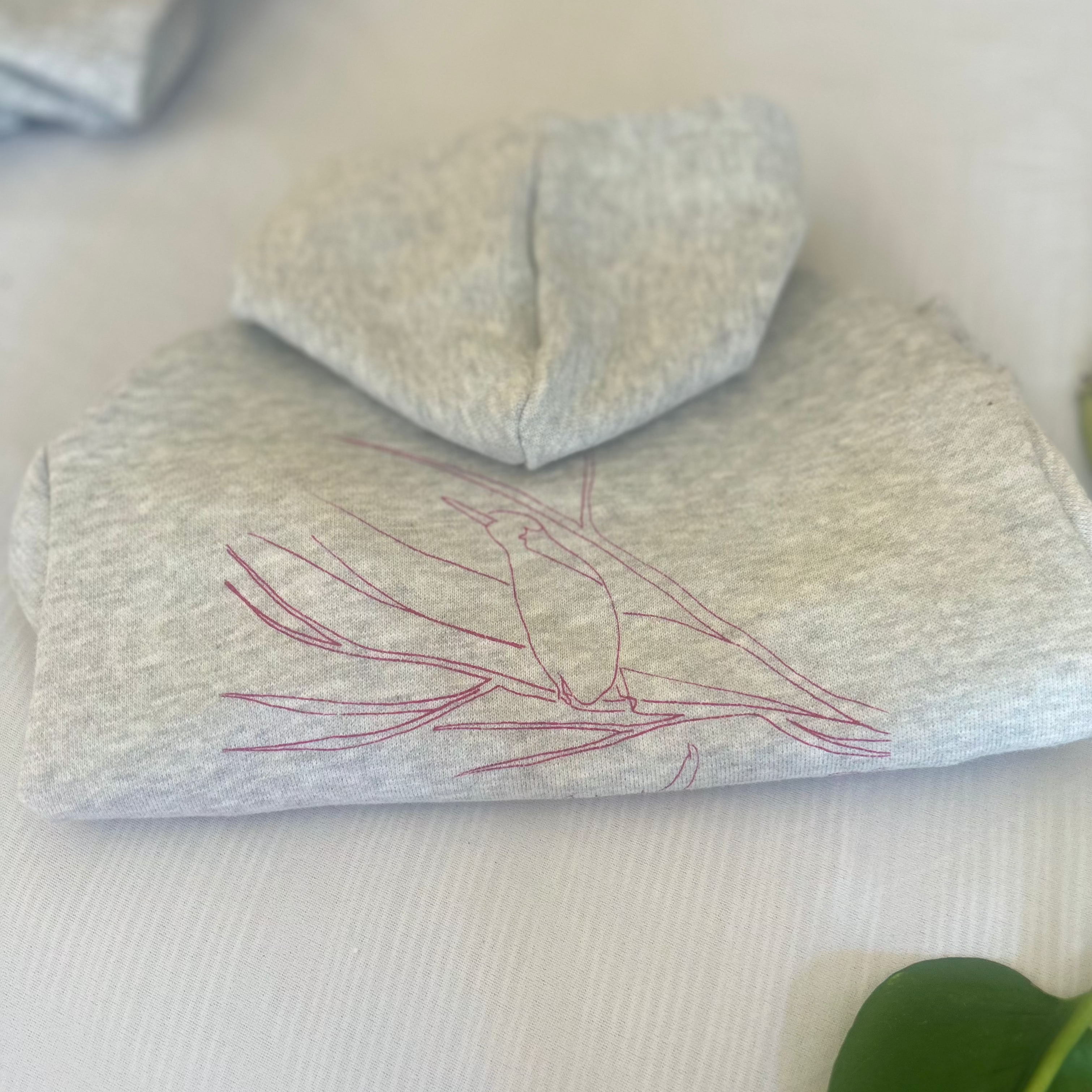 A neatly folded flat lay of the rose-printed ‘Charlie and the Bird’ hoodie in size small, showcasing the intricate bird design prominently. The arms are tucked in, and the hoodie lies flat on a white background with a subtle pinstripe texture, adding a touch of elegance. The corner of another hoodie is partially visible in the left, while a leaf is subtly cropped in the right corner, enhancing the natural feel of the image